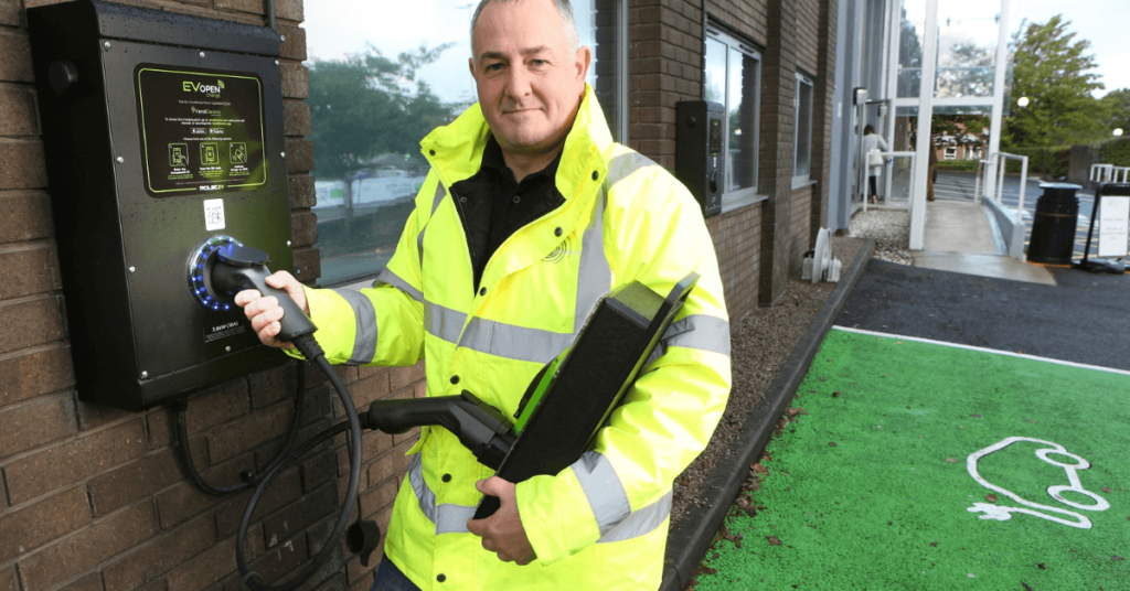 Antony Grace from ASH Integrated Services in a yellow jacket holding an electric car charger