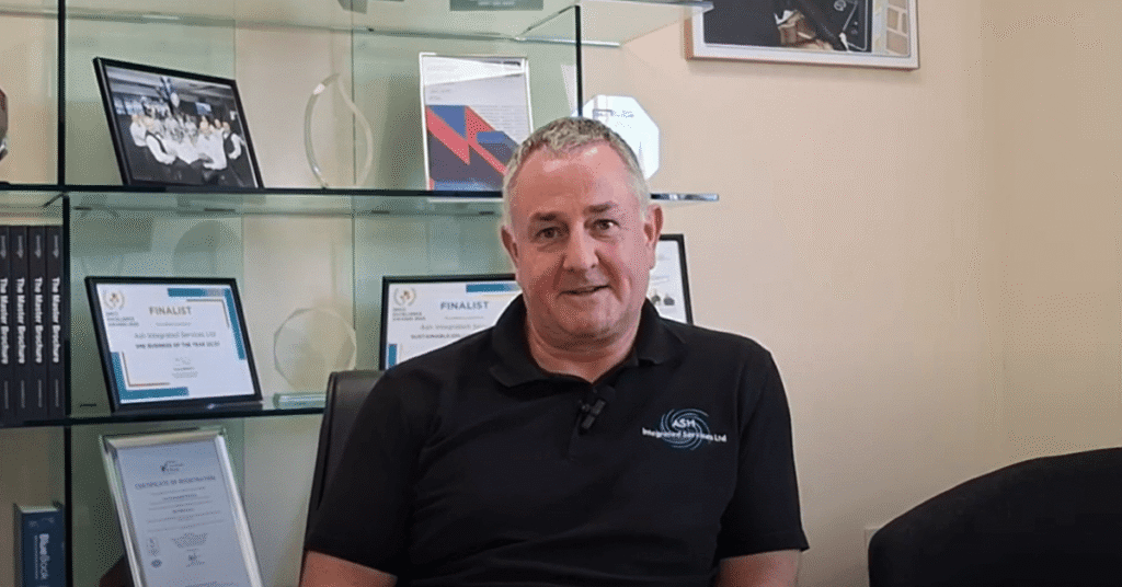 Image shows Antony Grace Director of ASH Integrated Services in front of glass shelving holding all their award certificates and trophies.