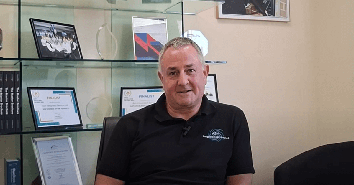 Image shows Antony Grace Director of ASH Integrated Services in front of glass shelving holding all their award certificates and trophies.