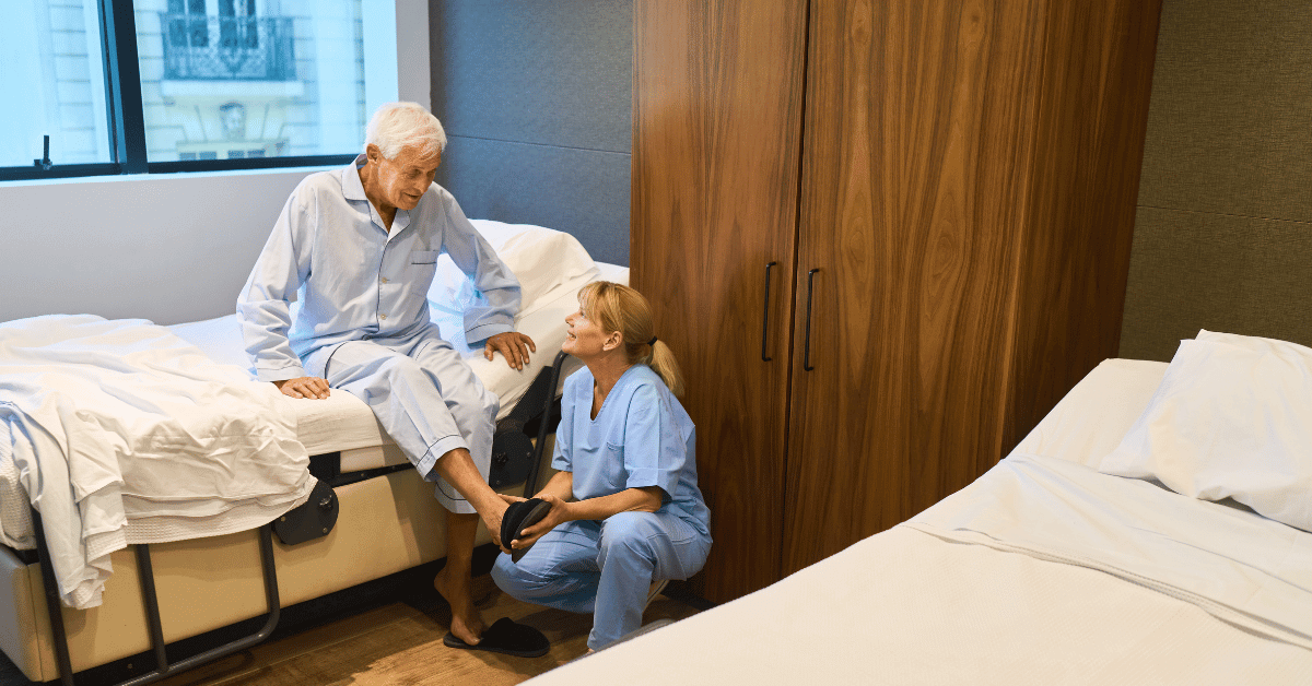 The picture shows a nurse crouching down, helping an older man to put his slippers on in a care home