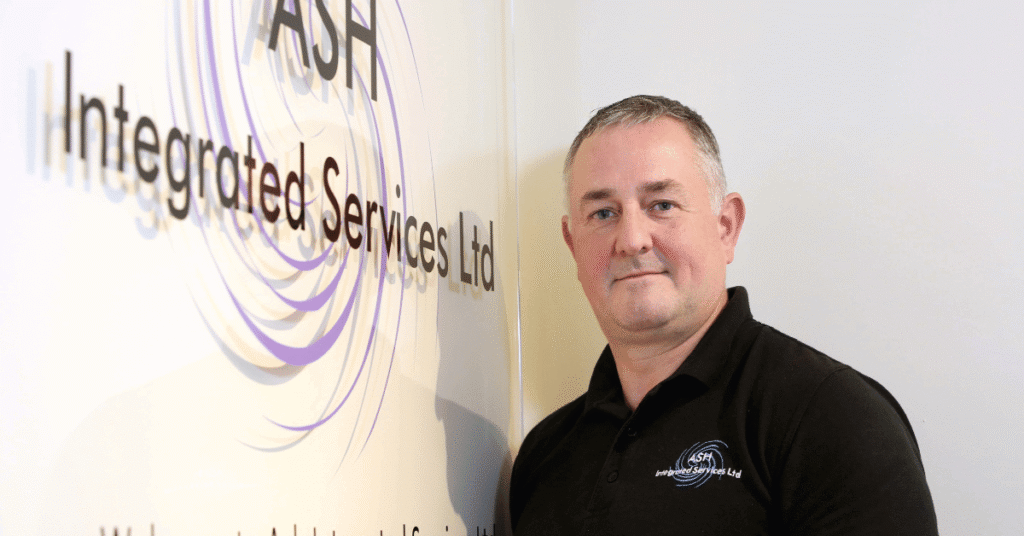 Image shows ASH Integrated Services Company Director Antony Grace stood in front of the company logo hung on a white wall