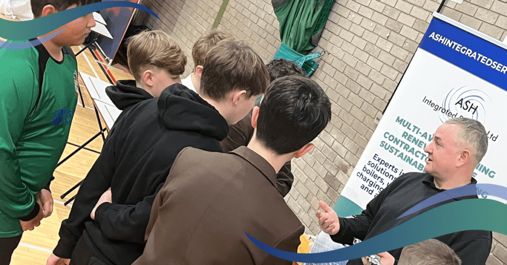 A man in a black shirt explains something to a group of young people in brown suits standing around a table with various items on it.
