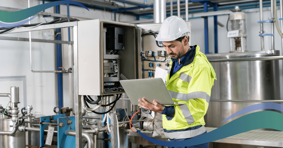 A technician wearing a hard hat and reflective vest inspects equipment in a factory while looking at a laptop.