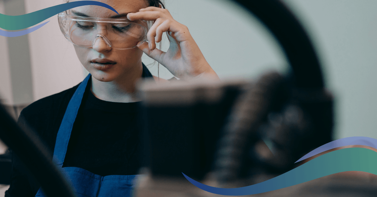 A person wearing safety glasses and a blue apron adjusts their glasses.