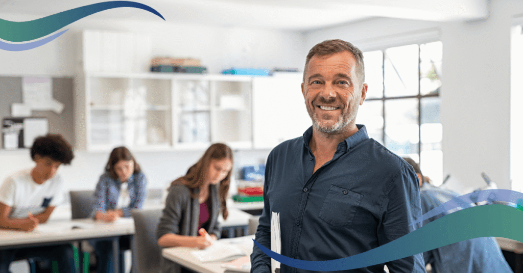 A smiling man in a blue shirt stands in a classroom with students working at desks in the background.