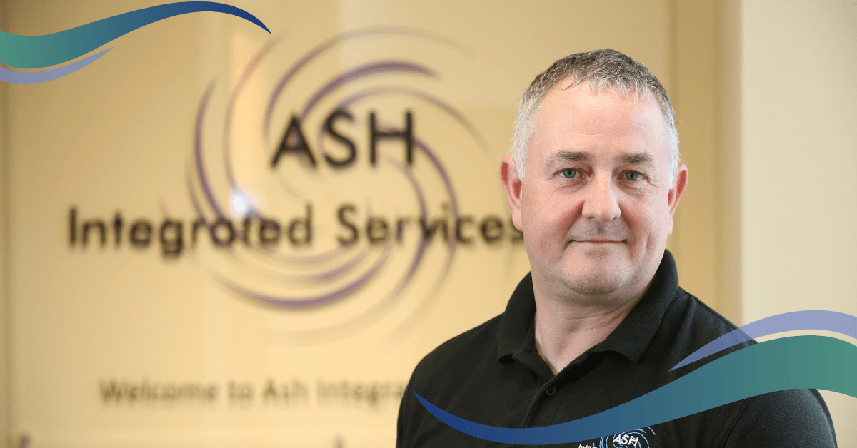 A man in a blue polo shirt stands in front of a glass sign that reads "ASH Integrated Services".