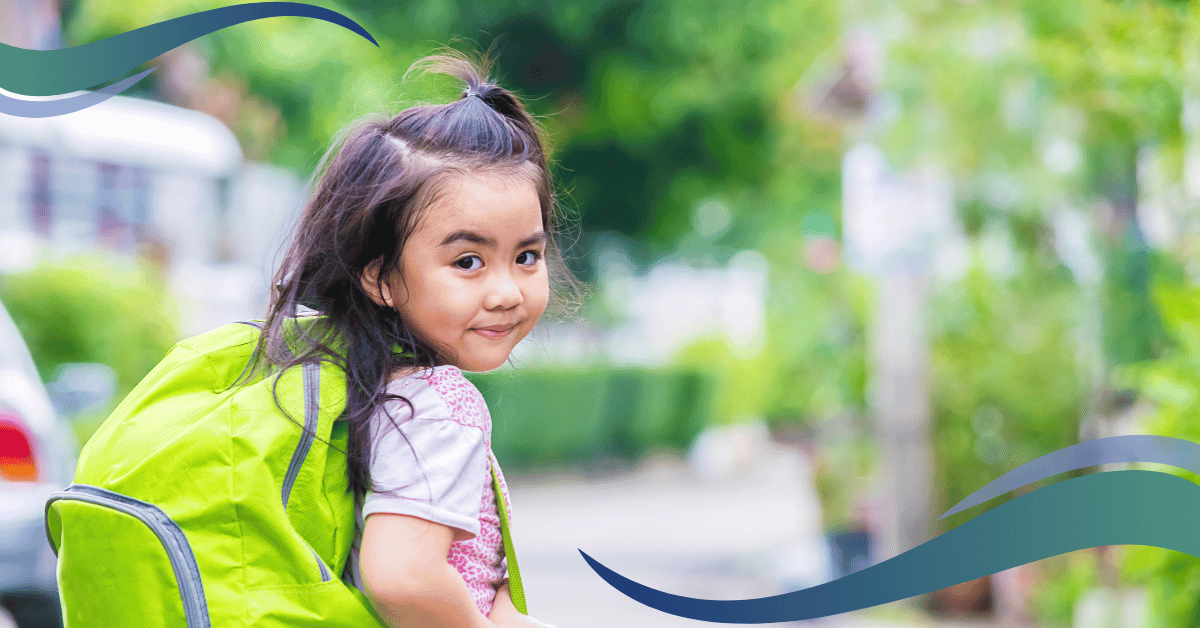 Image shows little girl looking over her shoulder smiling, with trees round her carrying a green backpack